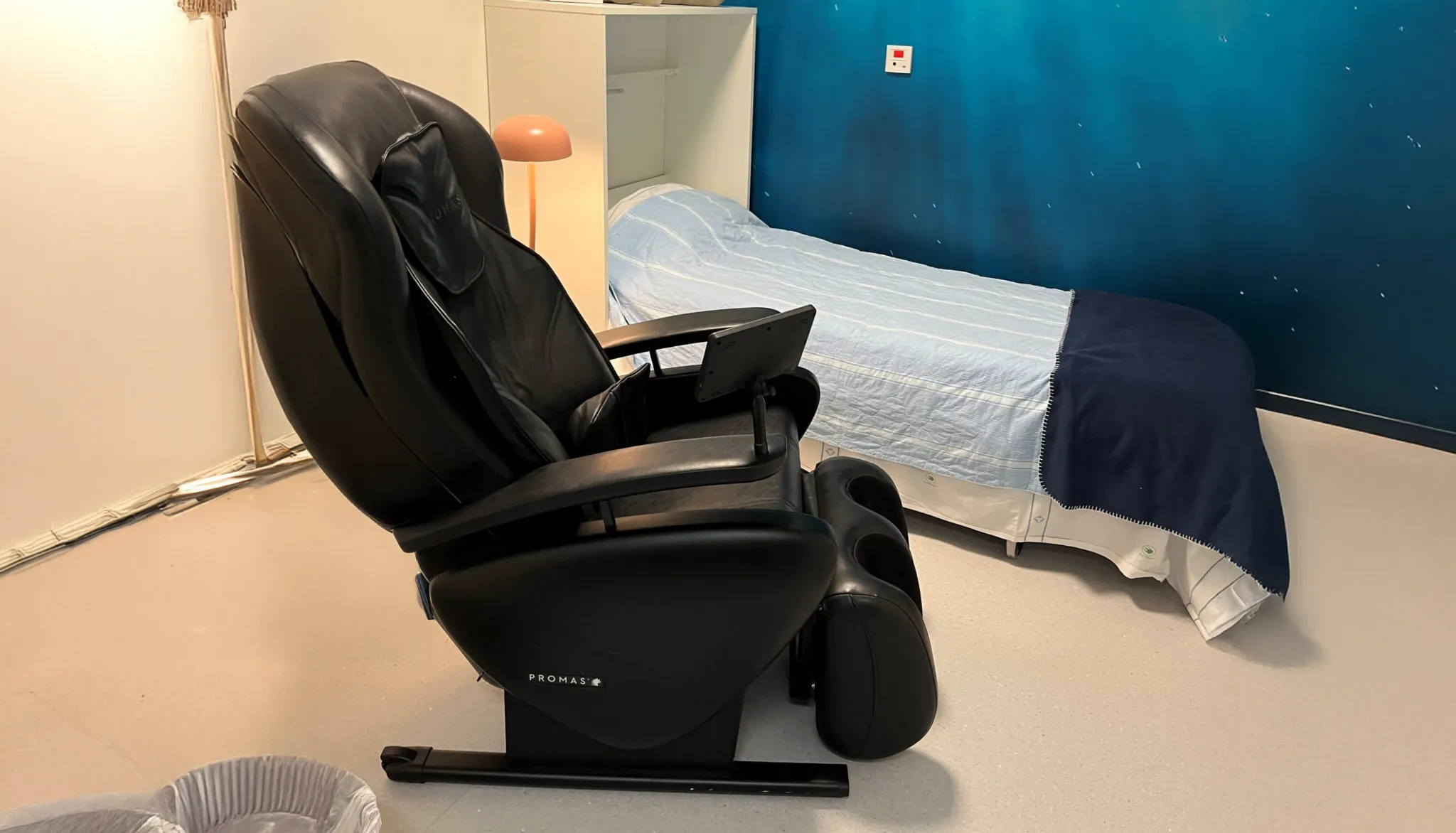Svart massagefåtölj bredvid en säng i ett rum med blågrön vägg